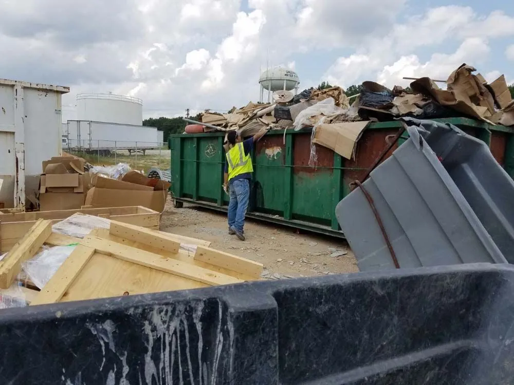 A man is standing in front of a dumpster filled with cardboard boxes.