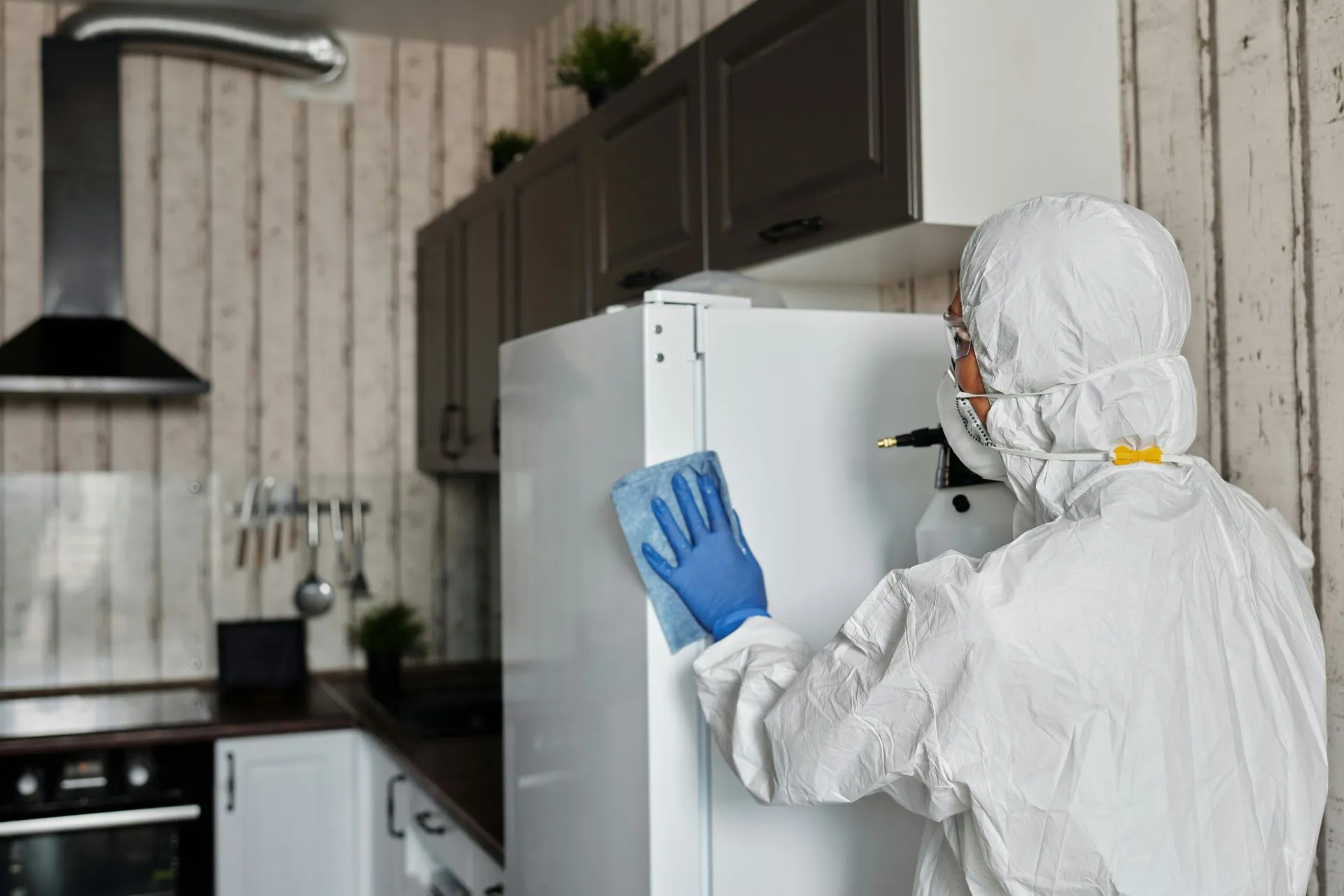 A person in a protective suit is cleaning a refrigerator in a kitchen.
