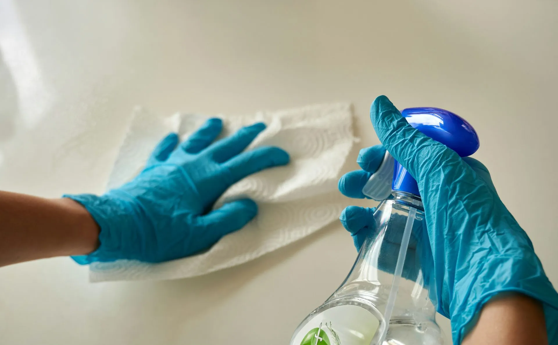 A person wearing blue gloves is cleaning a table with a towel and spray bottle.