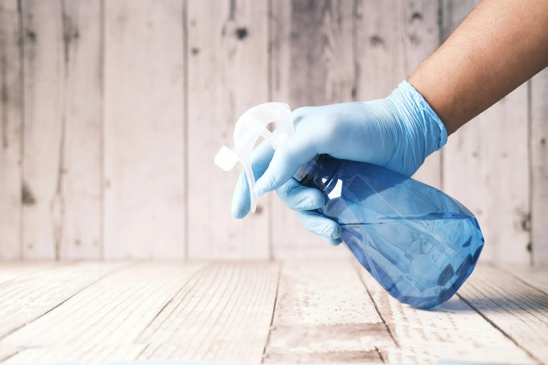 A person wearing blue gloves is holding a spray bottle on a wooden table.