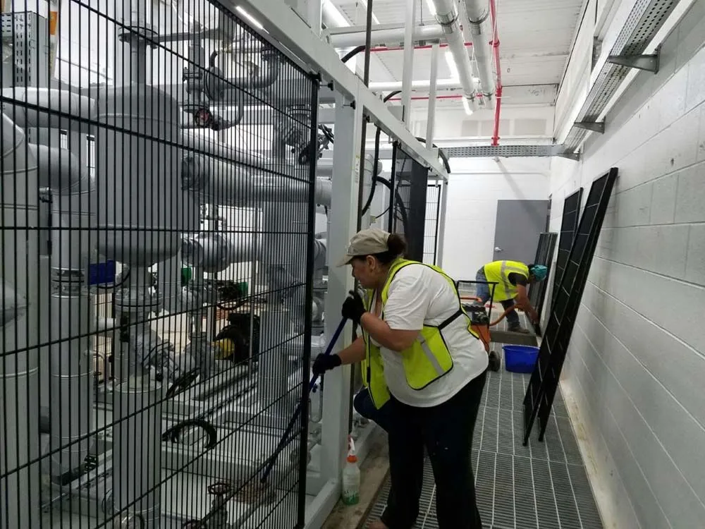 A woman in a yellow vest is cleaning a fence in a factory.